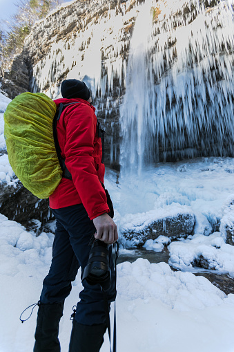 Photographer taking photos of frozen waterfall Pericnik in Slovenia. He is wearing a red jacket and a backpack. Winter time.