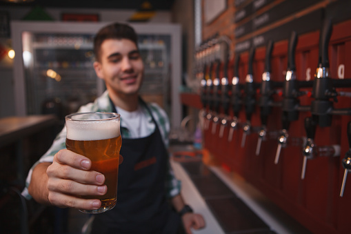 Happy bartender holding out beer glass to the camera, working at his bar