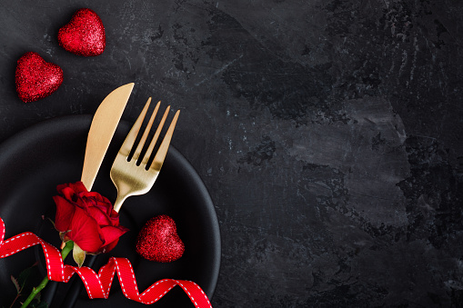 Valentine's Day table setting with plate, gold knife, fork, red rose, ribbon and hearts on dark background, top view, copy space. Valentines day background, holiday concept.