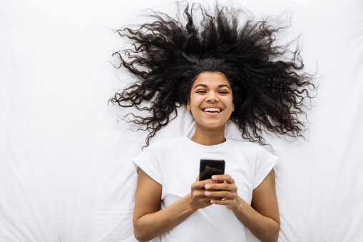 Chatting or shopping online. Pretty smiling african american woman lying on a white bed holding a smartphone and looking at the camera. Lazy morning