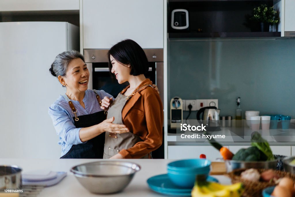Senior asian woman helping her daughter put on an apron before cooking Image of an Asian Chinese mother helping her daughter put on an apron and getting ready to cooking Senior Adult Stock Photo