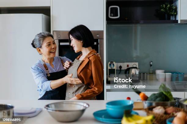 Senior Asian Woman Helping Her Daughter Put On An Apron Before Cooking Stock Photo - Download Image Now