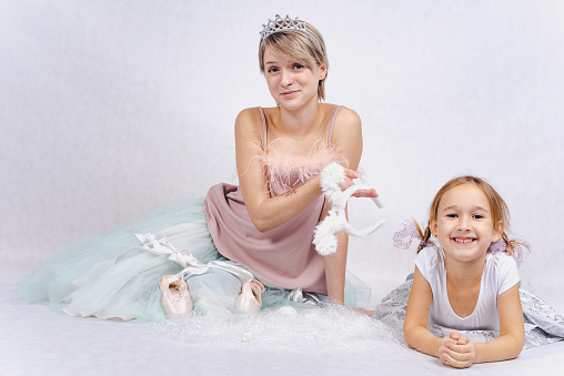 A cute little girl with two braids in a white dress with a young attractive mother in a pink T-shirt and a soft blue fluffy skirt is isolated on a white background. Girl with a crown on her head on a white background.