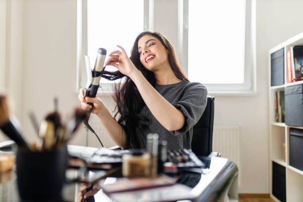 istock Woman looking at computer and using her curling iron while sitting at the dressing table. Woman does her hair by watching a tutorial on the internet at the time of the corona virus pandemic 1298155402