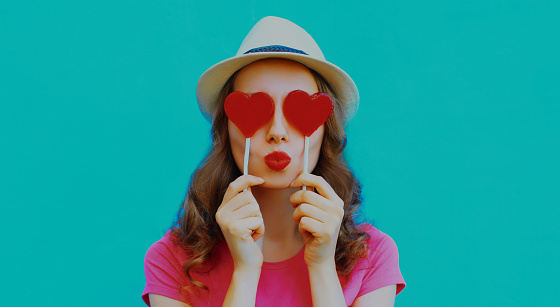 Portrait of young woman covering her eyes with red heart shaped lollipops on a blue background
