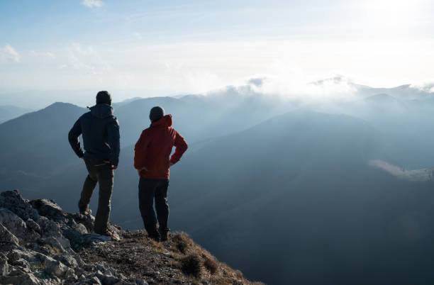 istock Two men standing standing with trekking poles on cliff edge and looking at sunset rays over the clouds. Successful summit concept image. 1298160745