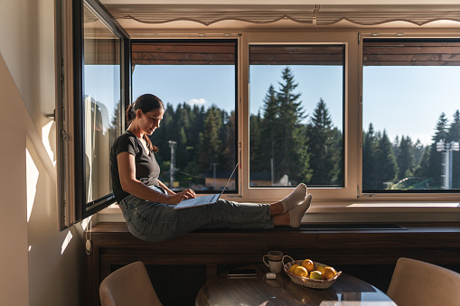 Young woman wearing headphones and sitting on window sill while working on laptop from home