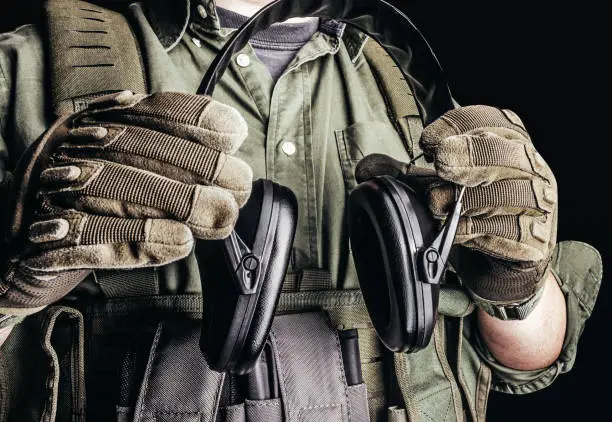 Photo of Soldier in tactical outfit with body armor, gloves and shirt holding shooting earmuffs.