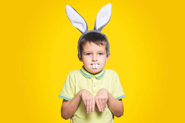 小さな男の子は黄色の背景に小さなイースターバニーとして機能しています。子供はイースターの日にバニーの耳と歯を身に着けています。 - easter egg easter yellow paint ストックフォトと画像