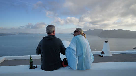 Santorini, Greece - 25 May, 2019: Senior couple travelers enjoy Santorini summer vacation sitting on wall having a beer drink and looking at amazing panoramic view over the roofs