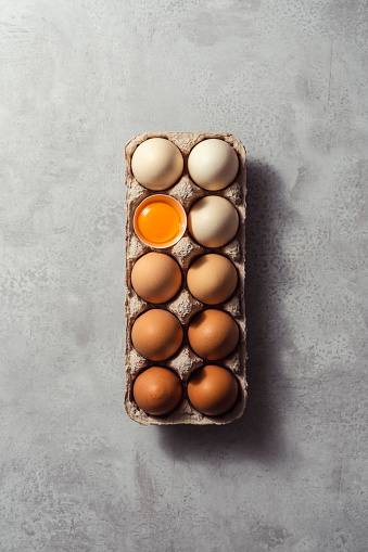 Box of color gradient brown eggs with one cracked egg, rectangular egg carton on grey background. Easter eggs. Shot from directly above.