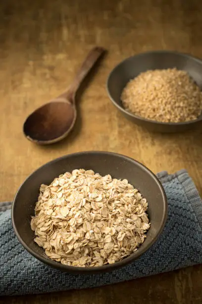 A close up of dried rolled oats and steel cut oats in bowls with a wooden spoon.