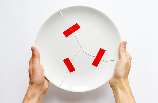 Top view of man hands holding a broken white plate, parts glued with red tape. Metaphor for divorce, relationships, friendships, crack in marriage. Love is gone. Isolated on white background.