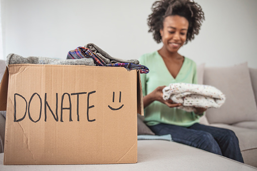 Happy African American young woman sit on couch stuck clothes in donation box at home, caring biracial female volunteer put apparel in carton package, donate to needy people, reuse, recycle concept