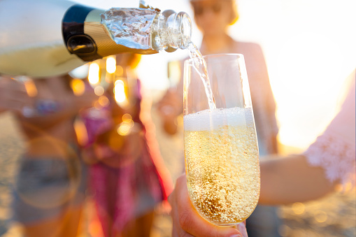 Pouring a glass of champagne with people partying in the background. The party is on the beach at sunset. Close up