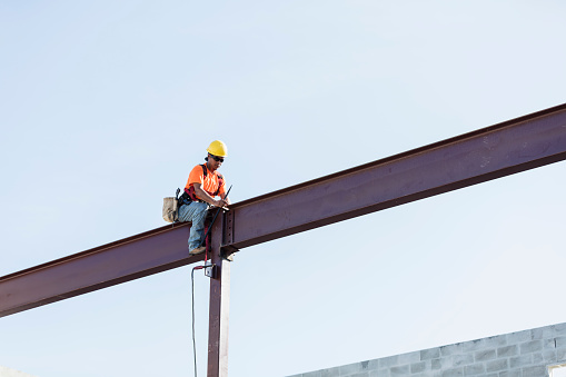 An Hispanic steel worker working high up on a girder. He is sitting on the girder, wearing a safety harness, working to secure the girder to a column.