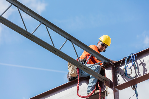 An Hispanic steel worker working high up on a girder. He is sitting on the girder, wearing a safety harness, working to secure a roof joist or truss to the girder.