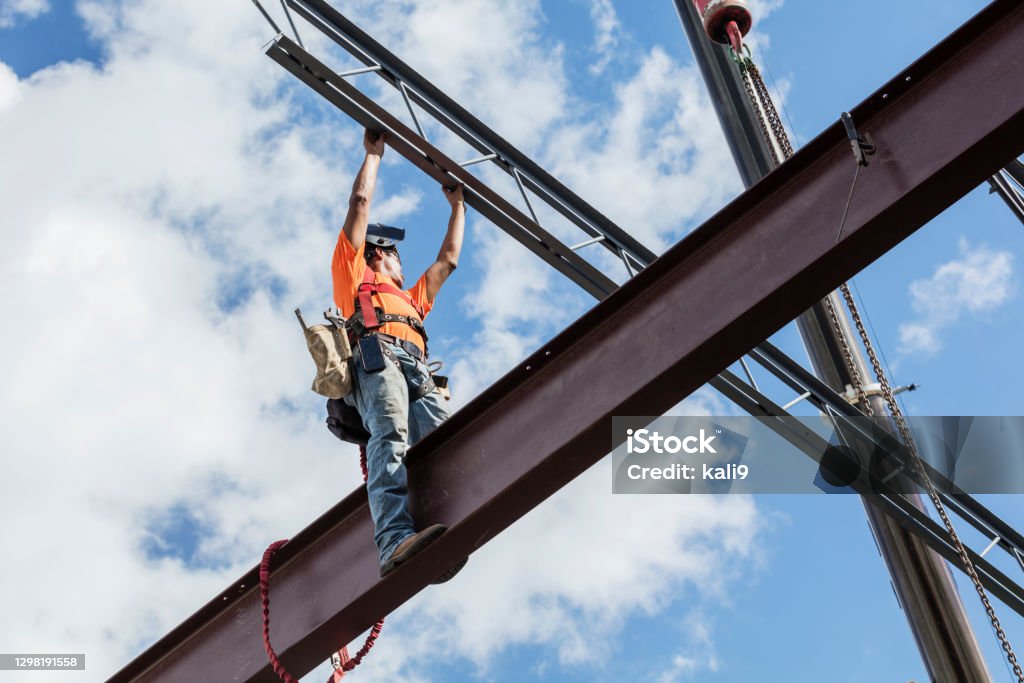 Ironworker at construction site installing roof joist An Hispanic steel worker working high up on a girder. He is standing on the girder, wearing a safety harness, reaching up for a roof joist or truss which is being lifted into place by a crane. He has a welding helmet on his head. Construction Site Stock Photo