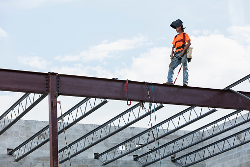 An Hispanic steel worker working high up on a girder. He is standing on the girder with wearing a welding helmet, working on attaching a roof joist to the girder.