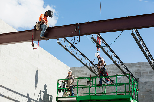 A multi-ethnic group of three workers at a construction site installing roof joists. Two of the workers are on a scissor lift and the third, an hispanic steel worker wearing a welding helmet, is on a girder working to secure the joist to it.