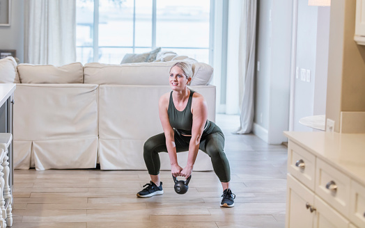 A mid adult woman in her 30s working out at home. She is in her living room doing squats while holding a kettlebell.