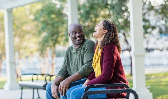 An African-American couple sit down on a park bench, smiling and laughing, having a good time. He is in his 40s and she is in her 30s. It is a cool, sunny, autumn day and they are enjoying each other's company.
