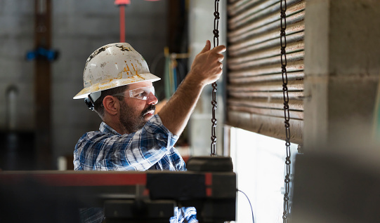 A mature man working in a warehouse, wearing a hardhat and safety goggles. He is pulling a chain to open the door of the loading dock.