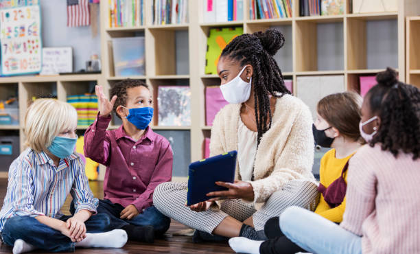 Preschool teacher, students in class, wearing masks A multi-ethnic group of kindergarten or first grade students and their teacher in the classroom, sitting together in a circle on the floor. The children are 5 to 7 years old. They are all wearing masks, back to school during the COVID-19 pandemic, trying to prevent the spread of coronavirus. The teacher, an African-American woman, is holding a digital tablet and looking at a boy who is raising his hand. teacher classroom child education stock pictures, royalty-free photos & images