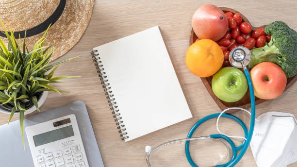 istock Notebook open blank note book paper page on work desk from top view with computer laptop, calculator, hat, healthy nutritious food  and safety KN95 medical face mask protection from PM2.5 and Covid-19 1298199142