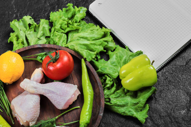 istock Raw chicken legs on wooden plate with fresh vegetables and open notebook 1298202307