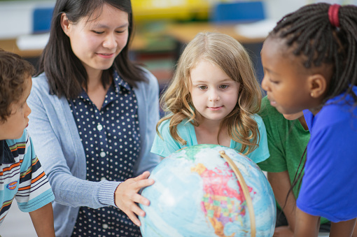 A female teacher of Asian ethnicity is showing her students a globe. They are all standing close to each other in class.