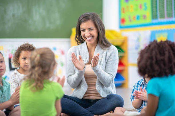 If you're happy and you know it, clap your hands A female teacher is sitting on the floor with her group of students at a classroom. They are all singing and clapping their hands together to a song. montessori education stock pictures, royalty-free photos & images