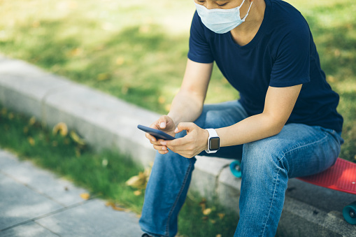 Asian woman wearing face mask sit on skateboard using smartphone in modern city