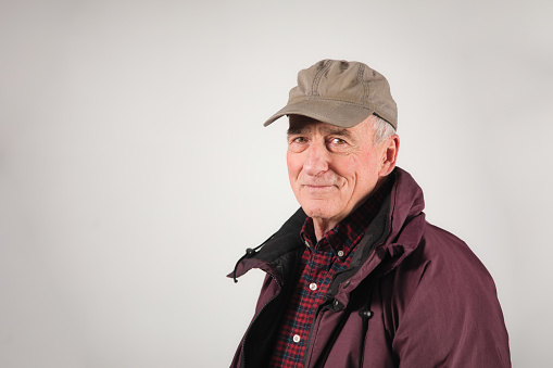 Senior man serene portrait in warm Winter clothing and wearing a baseball cap on a gray wall background.