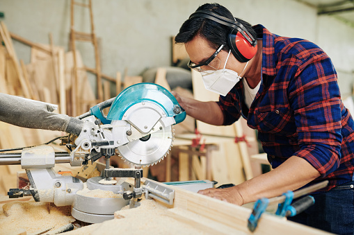Professional carpenter in protective mask, earmuffs and goggles cutting wooden planks with circular saw