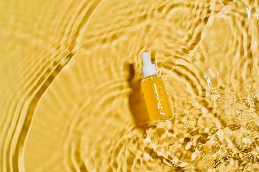 Bottle serum with vitamin C, cosmetic in clean transparent water with sunlight on yellow background, flat lay, top view, copy space