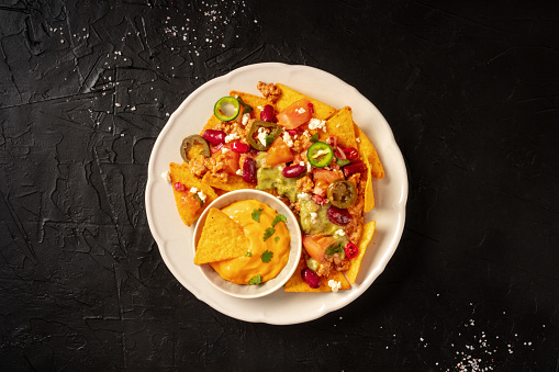 Mexican nachos with chili con carne, guacamole and cheese sauce, overhead shot on a black background