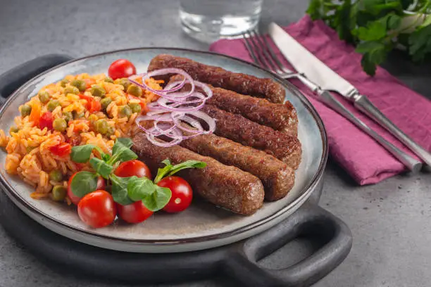 Balkan dinner with beef cevapcici and djuvec rice and vegetables, decorated with red onion. On grey table.
