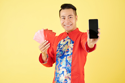 Cheerful young Asian man showing red lucky money envelopes and smartphone with blank empty screen