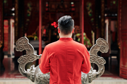 Young man in red shirt praying at copper urn in temple, view from the back