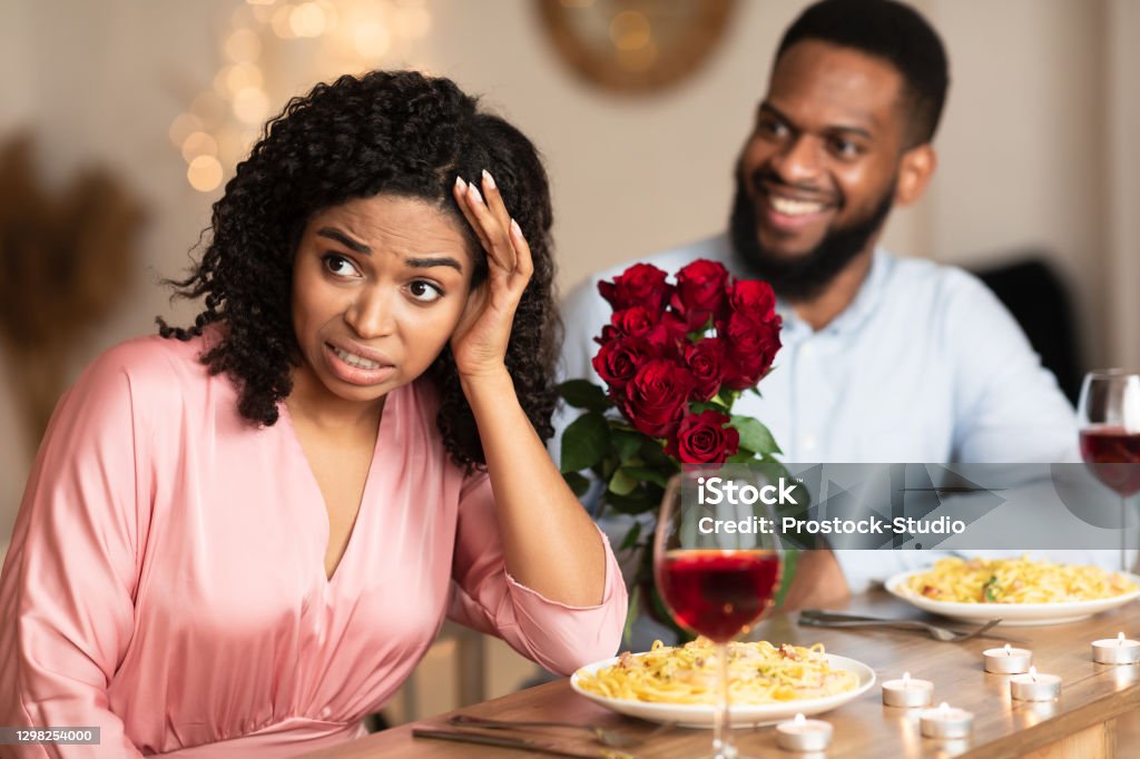 Black Woman On Unsuccessful First Date In Restaurant Bad First Impression And Blind Date Concept. Dissatisfied shocked black woman rejecting excited emotional obsessed man who giving her flowers, young couple sitting at table in cafe Dating Stock Photo
