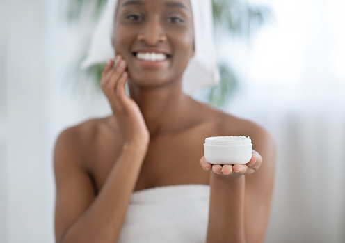 Moisturizing, lifting, nourishing day cream, soft skin. Smiling millennial african american lady in white towel, smears face with cream and holds jar in hand at home in bedroom interior in morning