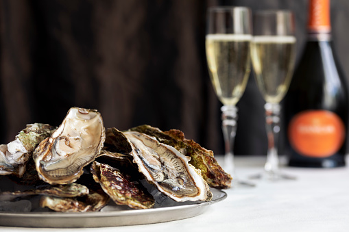 Plate of fresh opened oyster's shells on ice, two glasses of champagne and bottle on a white table. Selective focus