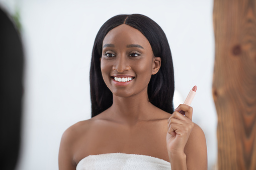 Modern cosmetics, makeup and beauty products. Smiling millennial african american lady with perfect skin looks in mirror and holds nude lipstick at home in bedroom interior in morning, empty space