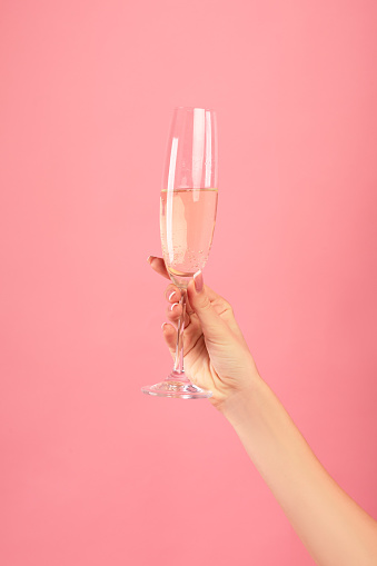 Closeup of female hand holding glass of champagne on pink studio background. Unrecognizable young lady having fun party, celebrating holiday with alcoholic drink, toasting and cheering