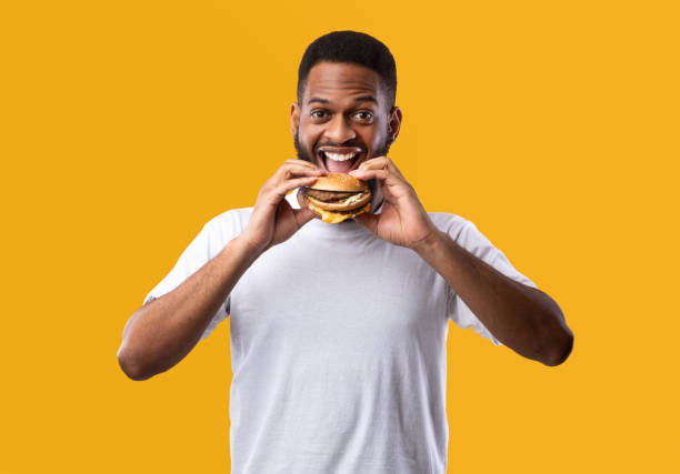 istock Joyful Hungry African Guy Eating Burger Standing On Yellow Background 1298255648