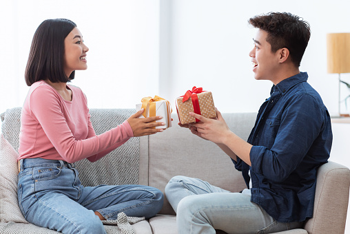 Valentine's Day Celebration. Lovely Asian Couple Giving Gifts Boxes Congratulating Each Other Celebrating Holiday Together Sitting On Sofa At Home. Romance, Holidays Presents Concept