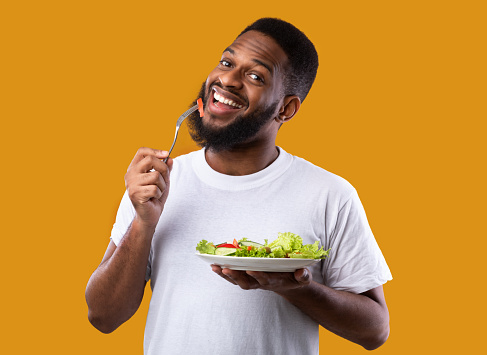 Cheerful Black Guy Eating Salad Posing Holding Plate And Fork Enjoying Healthy Meal Standing Over Yellow Studio Background, Smiling To Camera. Vegan Dinner Recipes, Weight Loss And Male Nutrition