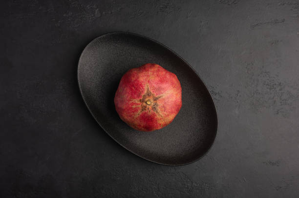 One pomegranate in the peel served in black oval plate on moody dark background. Top view, healthy diet concept, copy space