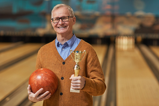 Waist up portrait of smiling senior man holding trophy and bowling ball while posing at bowling alley after winning match, copy space